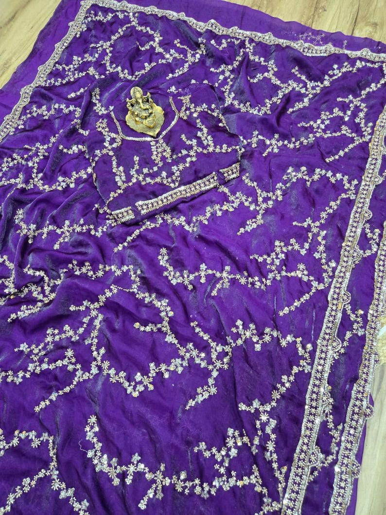May include: A vibrant purple saree with intricate silver embroidery. The fabric is draped, showcasing a flowing design. The saree features a detailed border and a decorative gold-colored centerpiece.