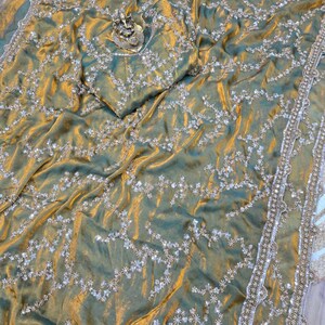May include: A shimmering, olive green sari with intricate silver embroidery and a matching blouse piece. The fabric has a subtle sheen, and the border is embellished with delicate beadwork. A small, ornate statue rests on the fabric.