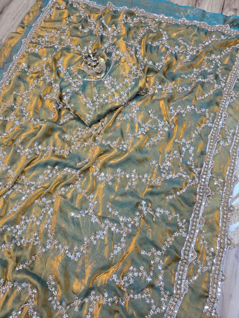 May include: A shimmering, olive green sari with intricate silver embroidery and a matching blouse piece. The fabric has a subtle sheen, and the border is embellished with delicate beadwork. A small, ornate statue rests on the fabric.