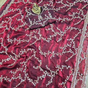 May include: A burgundy saree with intricate floral embroidery in silver and gold. The saree features a matching burgundy blouse piece with similar embellishments. The border is detailed with silver and red accents, creating a luxurious look.