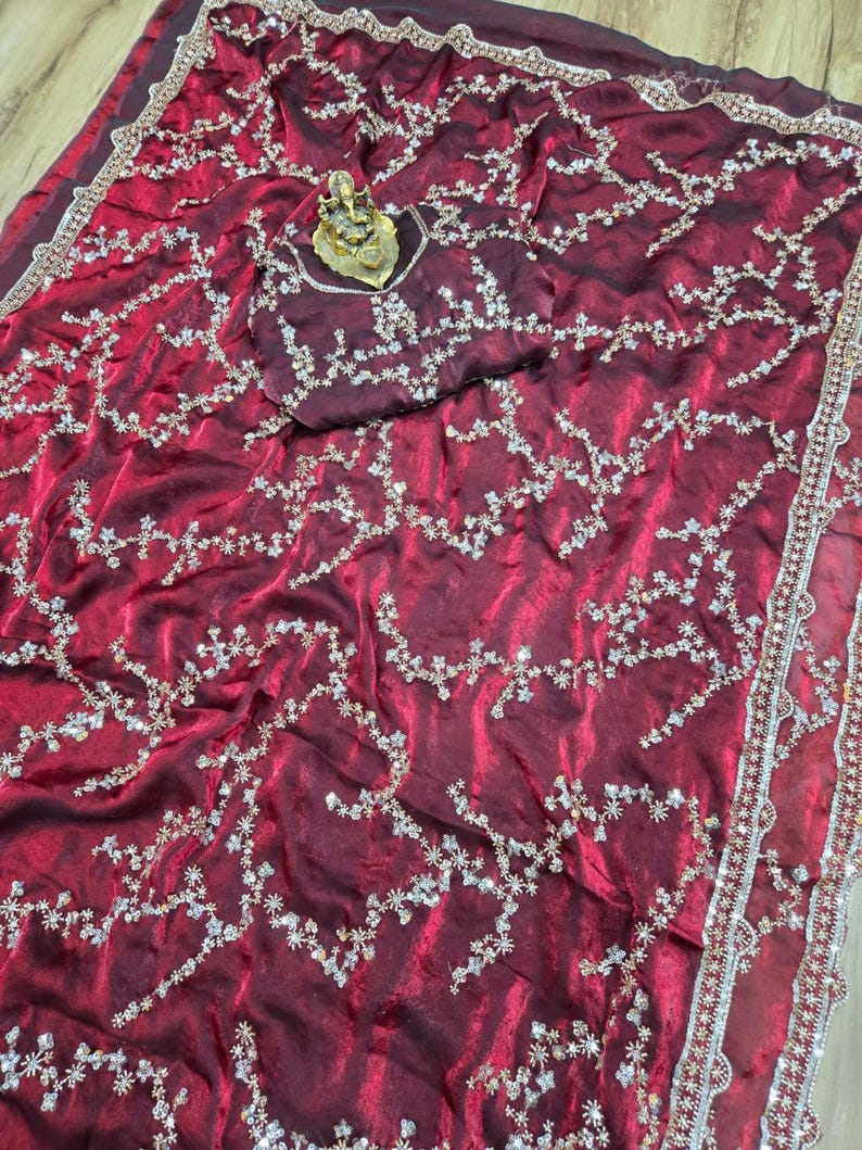 May include: A burgundy saree with intricate floral embroidery in silver and gold. The saree features a matching burgundy blouse piece with similar embellishments. The border is detailed with silver and red accents, creating a luxurious look.
