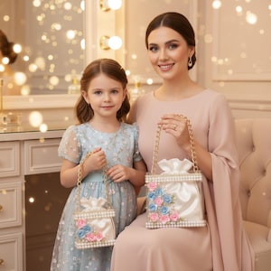 Könnte beinhalten: Zwei Frauen halten passende weiße Handtaschen mit goldenen Kettenriemen. Die Handtaschen sind mit rosa und blauen Stoffblumen und Perlenakzenten verziert. Die Frauen tragen Kleider, eines hellblau und das andere blassrosa.