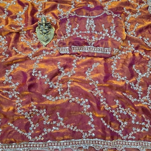 May include: A shimmering, rust-colored saree with intricate silver embroidery and a matching blouse. The fabric has a subtle sheen, and the design features floral patterns and a decorative border. A small, gold-colored statue is placed on the saree.