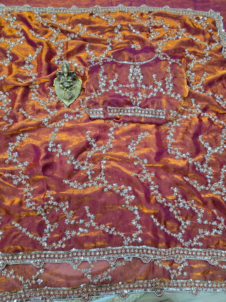May include: A shimmering, rust-colored saree with intricate silver embroidery and a matching blouse. The fabric has a subtle sheen, and the design features floral patterns and a decorative border. A small, gold-colored statue is placed on the saree.