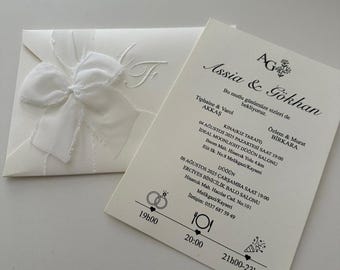May include: Ivory wedding invitation set. The invitation card features elegant script and event details. The envelope has a large, textured bow and embossed initials. The set is perfect for a wedding or special event.