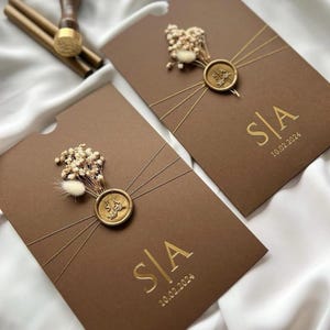 May include: Two brown invitation cards with gold foil lettering and a wax seal. Each card features the initials "S|A" and the date "10.02.2024". The cards are adorned with dried flowers and gold string.