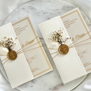 Op de afbeelding: Ivoor trouwuitnodigingen met gouden accenten, versierd met gedroogde bloemen en een zegel. De uitnodigingen zijn vastgebonden met touw en bevatten de namen "& Onur" en de datum 2024.