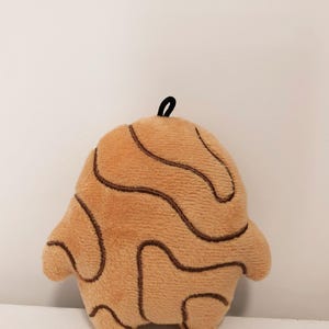 May include: A plush toy shaped like a rounded, abstract figure with a light tan, fuzzy texture. The surface features dark brown, embroidered, wavy, and curved lines that create a topographic pattern. A small black fabric loop is attached to the top.