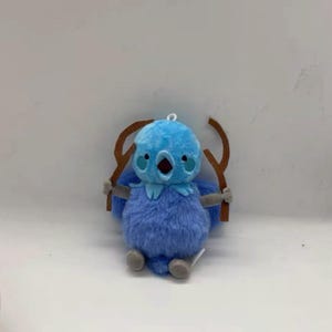 May include: A plush blue bird with a light blue head, open beak, and brown twig-like arms. The bird has grey feet and a small loop for hanging. The plush toy is designed to look like a cartoon character.