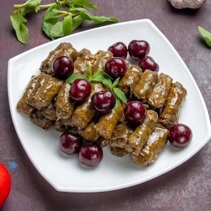 May include: A white square plate holds a serving of stuffed grape leaves, garnished with fresh cherries and mint. A red tomato, garlic cloves, and a lemon wedge are arranged around the plate on a dark purple surface.