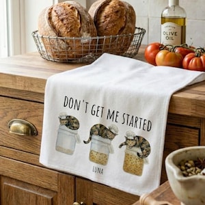 Puede incluir: Un paño de cocina blanco con el texto "DON'T GET ME STARTED" y tres ilustraciones de gatos atigrados con gorros de chef, cada uno sentado sobre un frasco de vidrio. Un gato está etiquetado como "LUNA". El paño está sobre una encimera de madera.