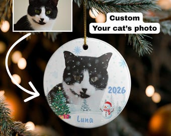 May include: A round white ceramic ornament featuring a custom black and white cat photo, the name Luna, the year 2026, a decorated Christmas tree, and a snowman. The text Custom Your cat's photo is displayed above the ornament hanging on a tree.