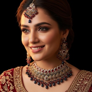 May include: A woman wearing a traditional Indian jewelry set, including a necklace, earrings, and a headpiece. The jewelry features red, blue, and gold accents. The woman is wearing a red and gold embroidered garment.