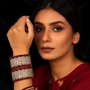 Puede incluir: Un par de brazaletes plateados adornados con filas alternas de piedras transparentes y rojo intenso. Los brazaletes están apilados en una muñeca, con un fondo oscuro. La modelo lleva una blusa roja.