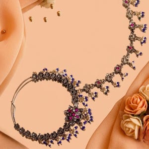 May include: An ornate silver-toned nose ring with a floral design. The jewelry features a large central flower with a magenta center and is adorned with small blue and white beads. The design extends in a curved line with additional floral elements and dangling beads.