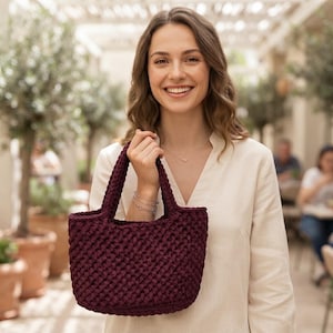 Pode incluir: Uma mala de mão em tom bordeaux profundo, confecionada com um padrão de crochet espesso e texturado. A mala apresenta duas alças integradas e um design estruturado com abertura superior, exibindo um ponto consistente e repetitivo.