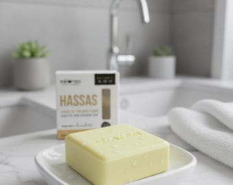 May include: A light yellow bar of soap in a white soap dish, with the word "inkaroo" embossed on the top. A box of "HASSAS" soap is behind it. The scene is set in a bathroom with a sink and a mirror.