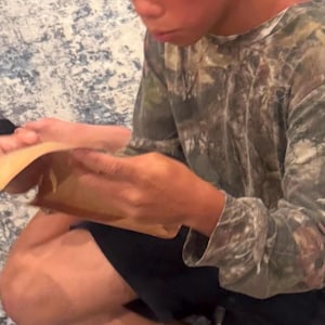 May include: A person wearing a camouflage long-sleeve shirt and black shorts. The person is holding a light brown object. The background is a patterned, light-colored surface.