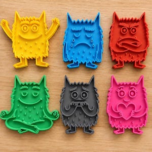 May include: A set of six colourful plastic cookie cutters shaped like monsters with various expressions. The collection includes a yellow happy monster, a blue crying monster, a red angry monster, a green meditating monster, a grey worried monster, and a pink monster holding a heart.