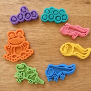 May include: A set of seven plastic biscuit cutters in various shapes and colours, including a purple cloud, a teal cloud, a pink tadpole, a yellow tadpole, a blue tadpole, a green lizard, and an orange frog, arranged in a circular pattern.