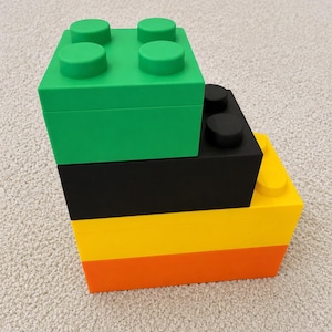 May include: A stack of four brightly coloured storage boxes, designed to look like large toy building blocks. The top box is green, followed by black, yellow, and orange. Each box has a flat top with circular studs.