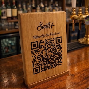 May include: A wooden table sign with the word "Swift" in cursive script and a QR code. Below the word "Swift" is the text "Follow Us On Facebook". The sign is on a wooden bar.