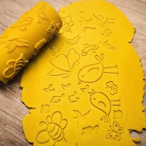 May include: A yellow cylindrical rolling pin with an embossed pattern of bees, birds, butterflies, and musical notes, positioned next to a flattened sheet of yellow dough that displays the same imprinted designs across its surface.