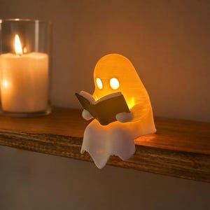 May include: A small, white, ghost-shaped lamp with a warm orange glow, depicted sitting and holding an open book. The ghost has two circular eye cutouts and is positioned next to two clear glass candle holders containing lit white pillar candles.