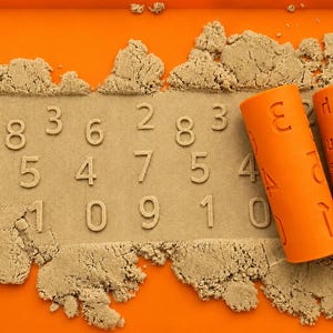 May include: Two orange cylindrical rollers with embossed numbers and letters rest on a bed of tan kinetic sand spread across an orange tray. The sand features imprinted numbers including 8, 3, 6, 2, 8, 3, 5, 4, 7, 5, 4, 1, 0, 9, 1, and 0.