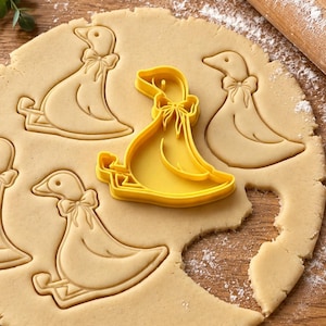 May include: A bright yellow plastic cookie cutter shaped like a goose wearing a bow tie, placed on a flat sheet of rolled-out beige cookie dough. The dough shows several impressed outlines of the same goose shape, creating a repeating pattern.