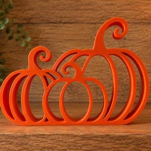 May include: A bright orange decorative cutout featuring three stylised pumpkins of varying sizes arranged side by side. The design consists of bold, rounded outlines with curved stems at the top, creating an open, hollow structure with smooth edges.