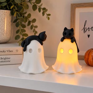 May include: Two white ghost-shaped lamps, each topped with a small black cat figurine. One ghost is unlit, while the other emits a warm, glowing yellow light from its hollow eyes and body. Both figures feature smooth, rounded surfaces and simple designs.