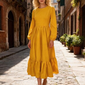 May include: A long-sleeved, mustard yellow dress with a round neckline and a tiered skirt. The dress features a fitted waist and a flowing, ruffled hem. The model is wearing white heels and is standing on a cobblestone street.