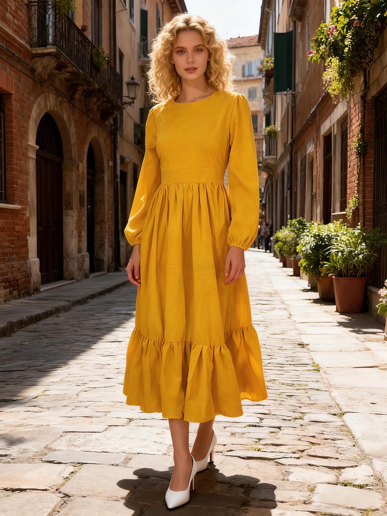 May include: A long-sleeved, mustard yellow dress with a round neckline and a tiered skirt. The dress features a fitted waist and a flowing, ruffled hem. The model is wearing white heels and is standing on a cobblestone street.