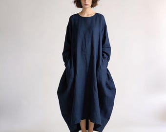 May include: A navy blue, long-sleeved dress with a loose, flowing silhouette. The dress features a round neckline and pockets. The hemline is slightly longer in the back. The model is wearing patterned flats.