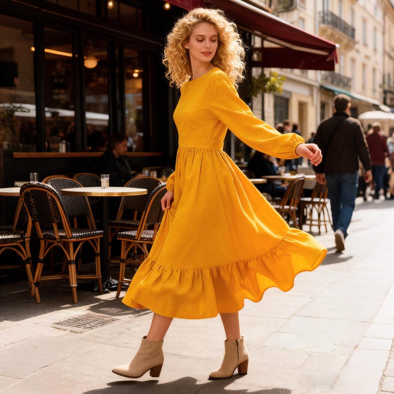 May include: A woman wearing a long-sleeved, mustard-yellow dress with a ruffled hem and beige ankle boots walks down a city street. The dress has a fitted bodice and a flowing skirt. The background includes a cafe with outdoor seating.