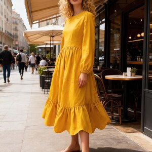 May include: A long, yellow dress with long sleeves and a tiered skirt. The dress has a round neckline and is worn with beige flats. The woman is standing on a sidewalk in front of a cafe.