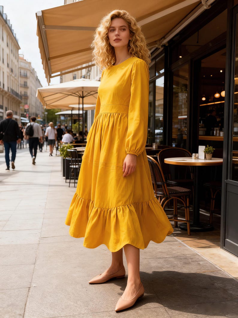 May include: A long, yellow dress with long sleeves and a tiered skirt. The dress has a round neckline and is worn with beige flats. The woman is standing on a sidewalk in front of a cafe.