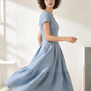 Pu&ograve; includere: Un abito azzurro con maniche corte e gonna a balze. L'abito ha uno scollo tondo e un corpetto aderente. La modella indossa l'abito in uno studio con luce naturale.