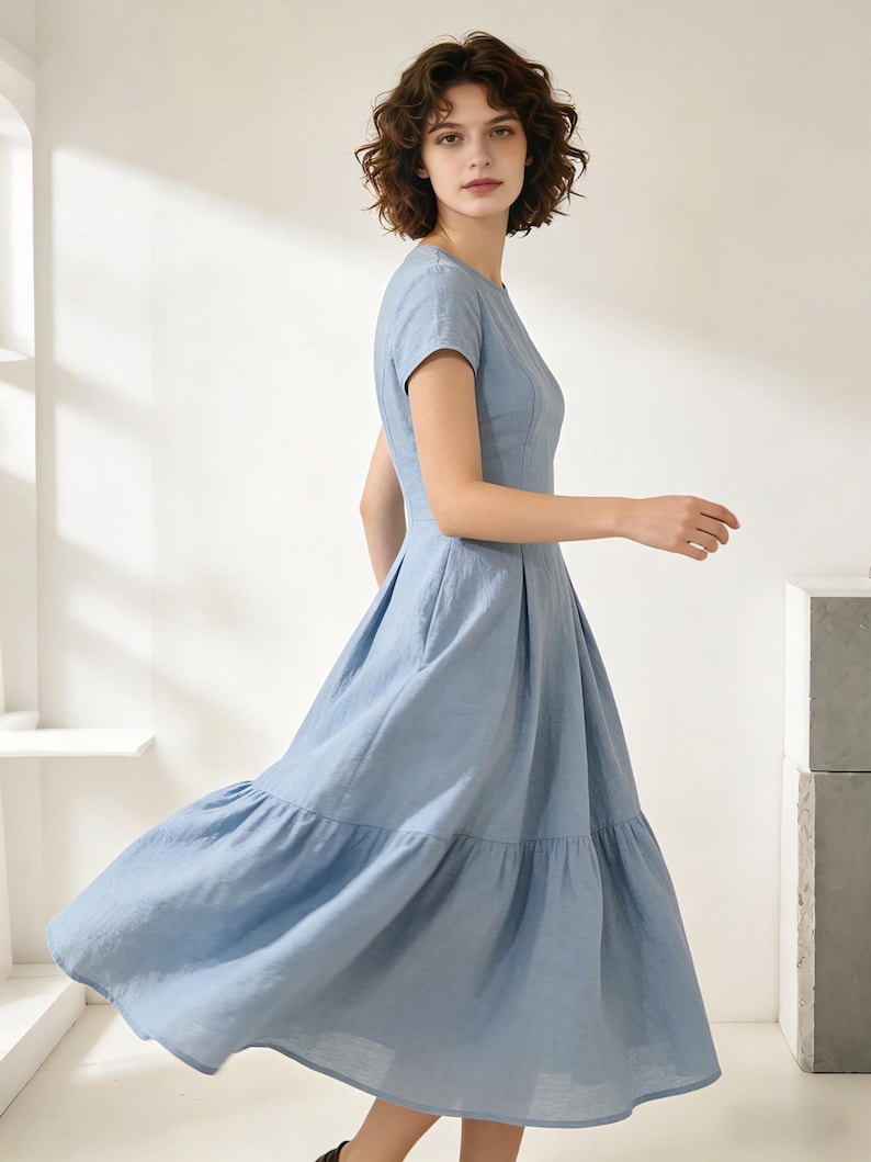 Pu&ograve; includere: Un abito azzurro con maniche corte e gonna a balze. L'abito ha uno scollo tondo e un corpetto aderente. La modella indossa l'abito in uno studio con luce naturale.