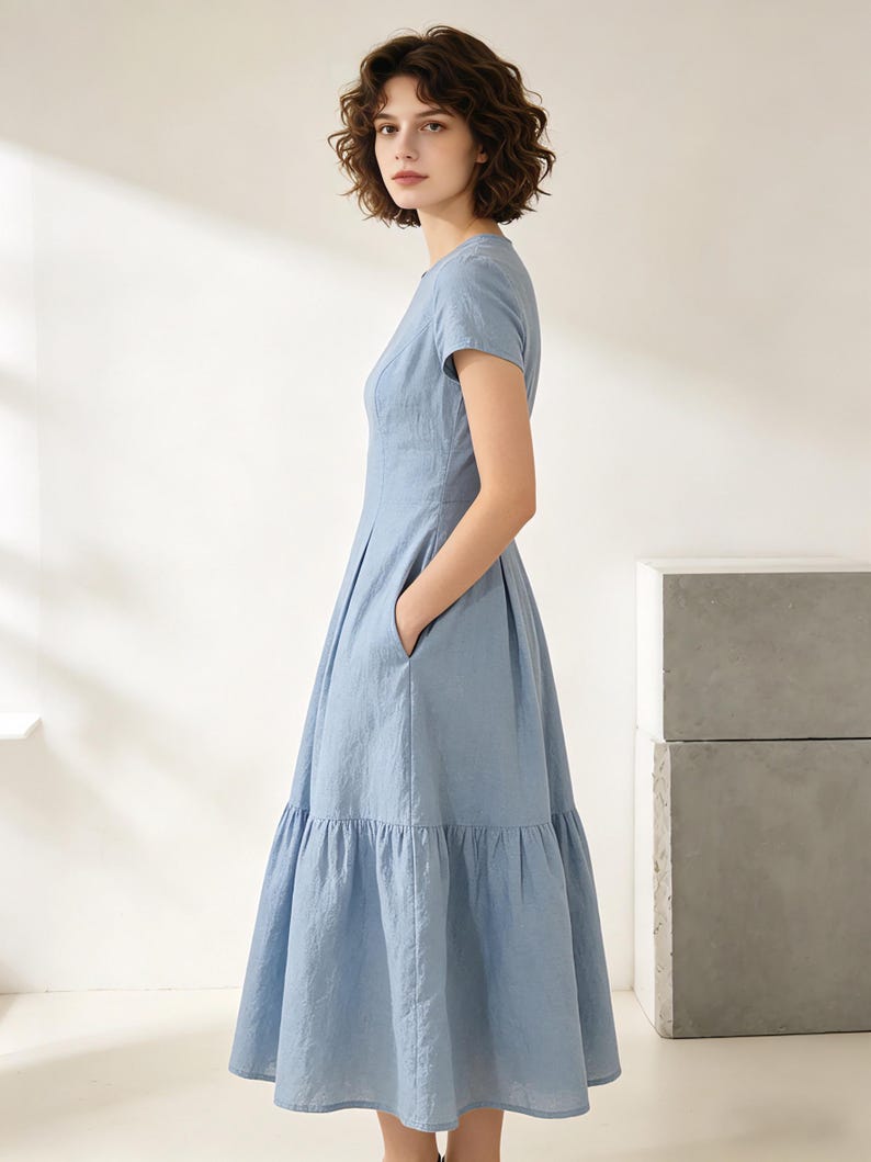 Pu&ograve; includere: Un abito azzurro con maniche corte e gonna a balze. L'abito presenta un corpetto aderente e tasche. La modella indossa l'abito in uno studio con sfondo neutro.