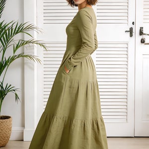 Op de afbeelding: Olijfgroene maxi-jurk met lange mouwen en een gelaagde rok. De jurk heeft een ronde hals en zakken. Het model draagt de jurk voor een witte deur met een plant in een gevlochten mand.