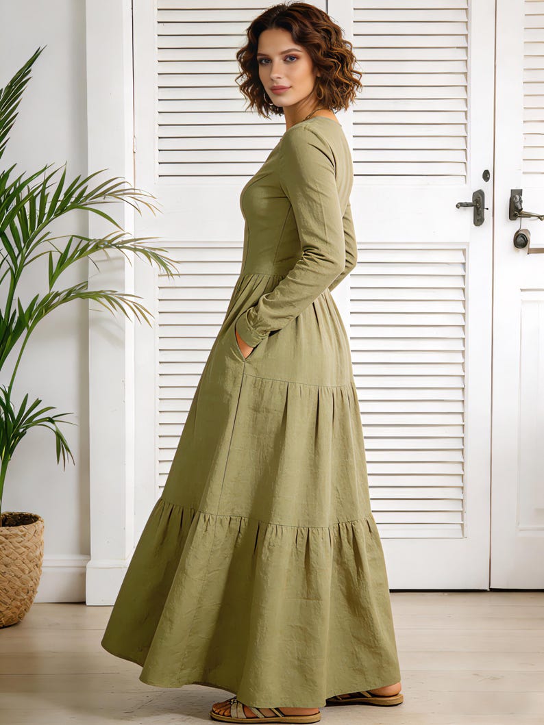 Op de afbeelding: Olijfgroene maxi-jurk met lange mouwen en een gelaagde rok. De jurk heeft een ronde hals en zakken. Het model draagt de jurk voor een witte deur met een plant in een gevlochten mand.