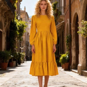 May include: A long-sleeved, mustard yellow dress with a round neckline, fitted waist, and a tiered, ruffled hem. The dress is paired with white high heels. The background is a cobblestone street lined with buildings and greenery.