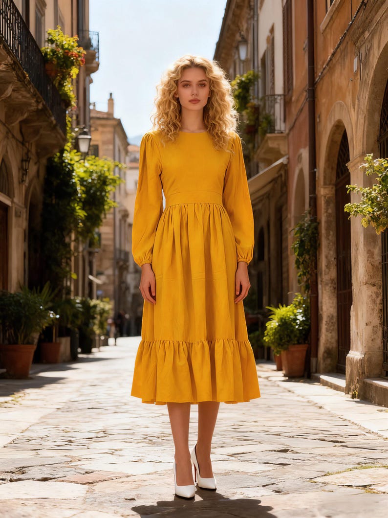 May include: A long-sleeved, mustard yellow dress with a round neckline, fitted waist, and a tiered, ruffled hem. The dress is paired with white high heels. The background is a cobblestone street lined with buildings and greenery.