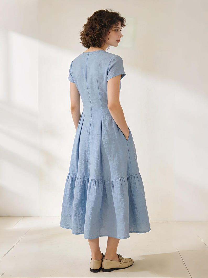 Pu&ograve; includere: Abito azzurro con maniche corte e gonna a balze. L'abito ha un corpetto aderente e una gonna svasata con tasche. La modella indossa scarpe beige. L'abito &egrave; realizzato in tessuto leggero.