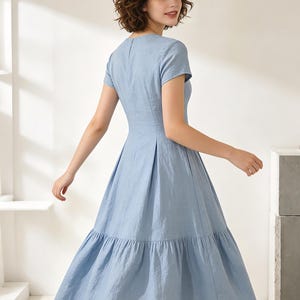 Pu&ograve; includere: Un abito azzurro con maniche corte e gonna a balze. L'abito ha un corpetto aderente e una gonna svasata, creando una silhouette classica. Il tessuto sembra essere un materiale leggero, adatto al clima caldo. La modella indossa l'abito.