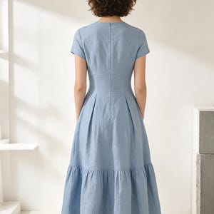 Pu&ograve; includere: Abito azzurro con maniche corte e gonna a balze. L'abito ha un corpetto aderente e una gonna svasata, con una sottile scollatura a V. Il tessuto sembra essere un misto leggero di lino o cotone.