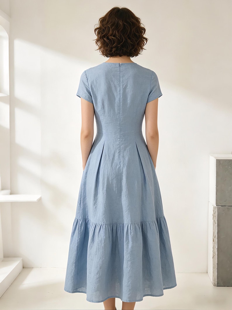 Pu&ograve; includere: Abito azzurro con maniche corte e gonna a balze. L'abito ha un corpetto aderente e una gonna svasata, con una sottile scollatura a V. Il tessuto sembra essere un misto leggero di lino o cotone.