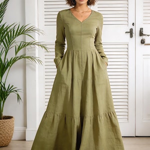 Op de afbeelding: Een olijfgroene maxi-jurk met lange mouwen en een V-hals. De jurk heeft een gelaagde rok, zakken en een getailleerd lijfje. Het model draagt sandalen. De achtergrond omvat een witte deur en een plant in een pot.